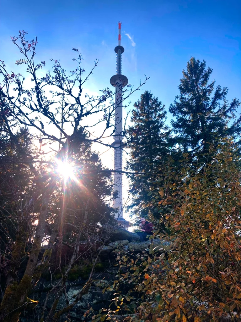 DG4KAI auf einem Fels in etwas Entfernung beim Funken auf dem Ochsenkopf. Im Hintergrund Sendemast BR Bayerischer Rundfunk. Sonnenlicht blitzt durch die Bäume in Richtung Fotograf